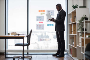 a black man standing in front of a window in an office using a laptop