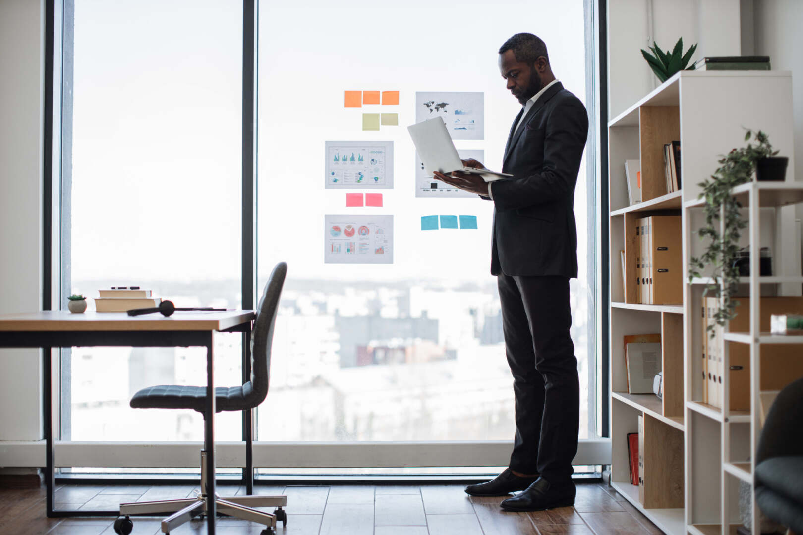 a black man standing in front of a window in an office using a laptop