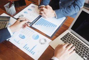 two people's hands working on a computer and a spreadsheet on a desk